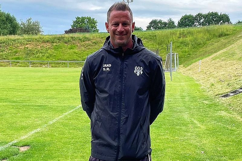Oberkörper-Portrait-Foto: Mann in schwarzer Trainingsjacke am Rande eines Sportplatzes